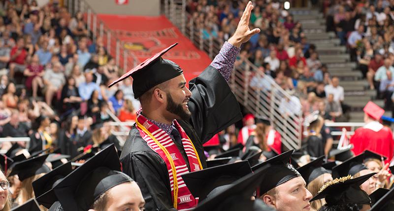Male in cap and gown standing in arena, waving at crowd.